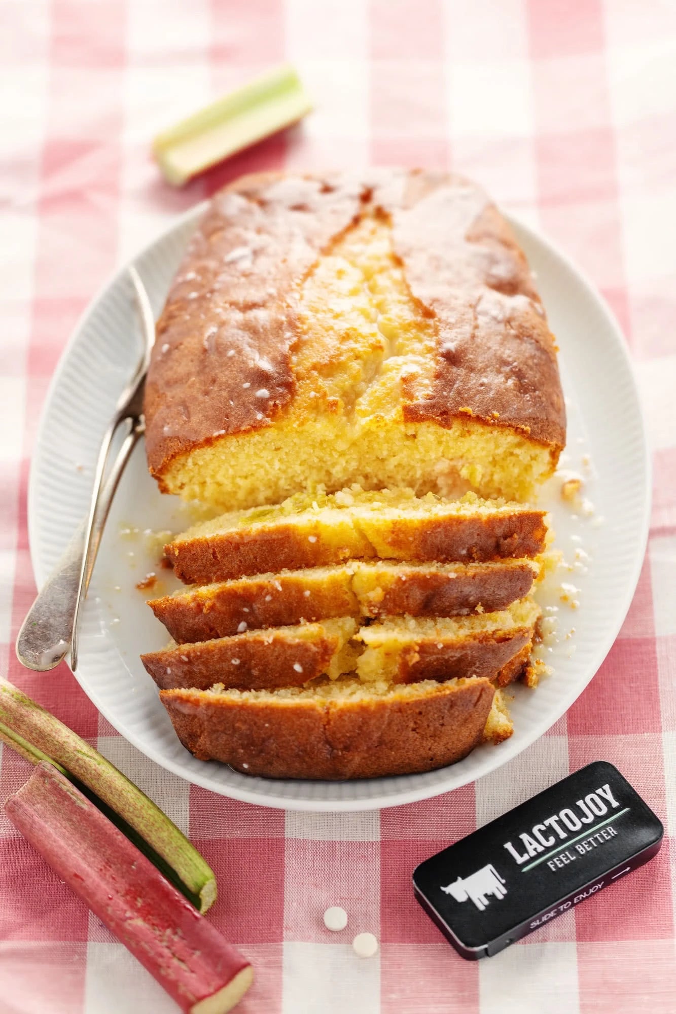 Loaf of cake sliced on a plate with rhubarb and LactoJoy package on a checkered tablecloth
